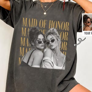 Könnte beinhalten: Ein anthrazitfarbenes Oversize-T-Shirt mit einer schwarz-weißen Grafik von zwei Personen mit Sonnenbrillen. Der Text "MAID OF HONOR" ist in großen goldenen Buchstaben hinter den Figuren gedruckt. Ein kleiner Einsatz zeigt das Originalfoto.