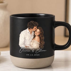May include: A black ceramic mug with a matte finish and a light beige base. The front features a printed illustration of a couple embracing, with the text "Olivia & Ethan" in a white script font and the date "09.14.2026" printed below in white.