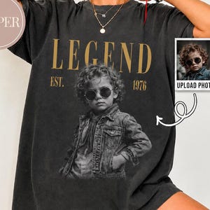 Könnte beinhalten: Ein anthrazitfarbenes Kurzarm-T-Shirt mit einer großen schwarz-weißen Grafik eines Kindes mit Sonnenbrille und Jeansjacke. Der Text LEGEND und EST. 1976 ist in Gold über und unter der Grafik gedruckt. Ein kleines Inset-Feld zeigt das Originalfoto.