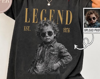 May include: A charcoal gray short-sleeve t-shirt featuring a large black and white graphic of a child wearing sunglasses and a denim jacket. The text LEGEND and EST. 1976 are printed in gold above and below the graphic. A small inset box shows the original photo.