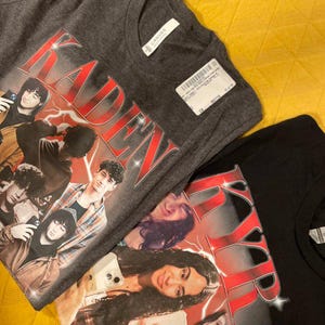 Puede incluir: Se muestran dos camisetas, una gris antracita y otra negra. La camiseta gris presenta un gr&aacute;fico con el nombre "KADEN" en texto rojo estilizado y un collage de personas. La camiseta negra tiene un dise&ntilde;o similar.