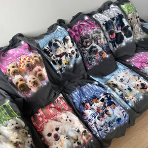 May include: A collection of dark gray t-shirts featuring various dog breeds in colorful, artistic designs. Each shirt showcases a different group of dogs, with text accents in pink, blue, and white. The shirts are displayed in a casual arrangement.