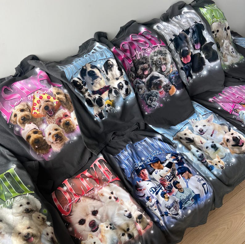 May include: A collection of dark gray t-shirts featuring various dog breeds in colorful, artistic designs. Each shirt showcases a different group of dogs, with text accents in pink, blue, and white. The shirts are displayed in a casual arrangement.