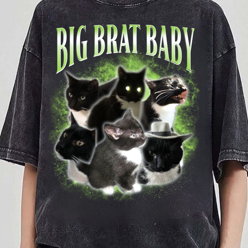 Puede incluir: Camiseta gris oscuro con un gr&aacute;fico de seis gatos blancos y negros con diversas expresiones, rodeados por un dise&ntilde;o verde moteado. El texto "BIG BRAT BABY" est&aacute; arqueado sobre los gatos en verde.