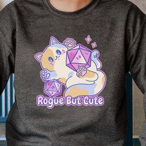 Può includere: Felpa grigia melange scuro con un gatto dei cartoni animati che tiene un dado viola e rosa con il testo "Rogue But Cute".