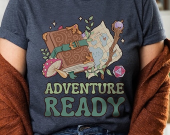 Puede incluir: Una camiseta azul marino con un gráfico de un libro, un mapa y dados. El texto "Adventure Ready" está impreso en verde en la camiseta.