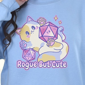 May include: Light blue sweatshirt with a cartoon cat holding a pink and purple 20-sided die. The cat is surrounded by other dice and the text "Rogue But Cute" is printed below the cat.