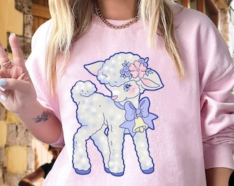 Peut inclure: Un sweat-shirt rose pâle avec un agneau de dessin animé portant un nœud bleu et une cloche. L'agneau porte une couronne florale sur la tête.