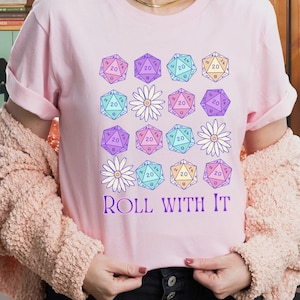 Puede incluir: Una camiseta rosa con un gr&aacute;fico de dados de 20 caras de color pastel y margaritas. El texto "ROLL WITH IT" est&aacute; impreso debajo del gr&aacute;fico.