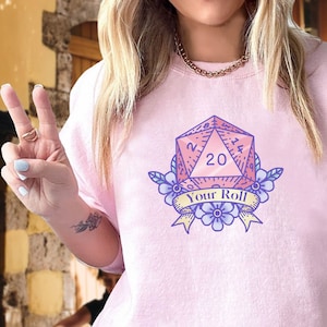 Puede incluir: Una sudadera rosa claro con un gráfico de un dado de veinte caras con flores y una pancarta que dice "Your Roll".