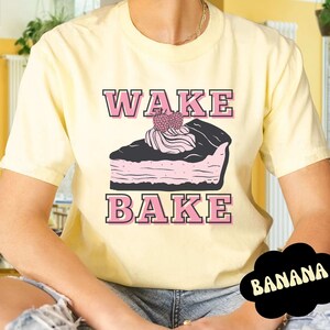 Puede incluir: Una camiseta amarilla con el texto "WAKE BAKE" en letras rosas. Una rebanada de pastel de chocolate de dibujos animados con glaseado rosa y una fresa en la parte superior está en el centro de la camiseta.