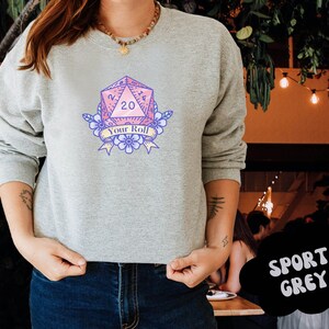 May include: A grey sweatshirt with a pink and blue graphic of a twenty-sided die with the text "Your Roll".