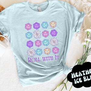 Puede incluir: Una camiseta azul claro con un dise&ntilde;o gr&aacute;fico de dados y flores. El texto "ROLL WITH IT" est&aacute; impreso en la camiseta.
