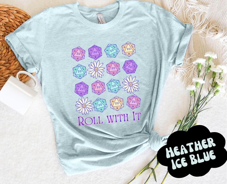 Puede incluir: Una camiseta azul claro con un dise&ntilde;o gr&aacute;fico de dados y flores. El texto "ROLL WITH IT" est&aacute; impreso en la camiseta.