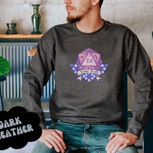 May include: A dark heather grey sweatshirt featuring a pink and purple 20-sided die graphic. The die is surrounded by flowers and a banner that reads "Your Roll".