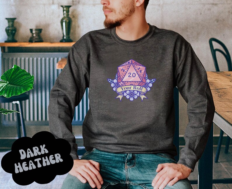 May include: A dark heather grey sweatshirt featuring a pink and purple 20-sided die graphic. The die is surrounded by flowers and a banner that reads "Your Roll".