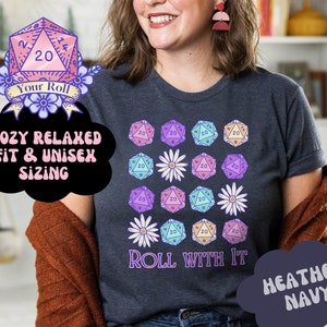 Puede incluir: Una camiseta azul marino jaspeada con un gr&aacute;fico de dados de 20 caras coloridos y margaritas blancas. El texto "Roll With It" est&aacute; impreso debajo de los dados. La camiseta tiene un ajuste c&oacute;modo y relajado y es de talla unisex.