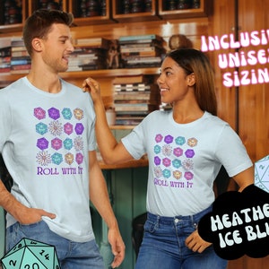 Puede incluir: Dos personas llevan camisetas de color azul claro con un dise&ntilde;o de dados coloridos y flores. El texto "Roll with it" est&aacute; impreso en las camisetas. Las camisetas est&aacute;n disponibles en Heather Ice Blue.