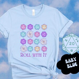 Puede incluir: Una camiseta azul claro con un dise&ntilde;o de varios dados de veinte caras de color pastel y margaritas blancas. El texto "ROLL WITH IT" est&aacute; impreso debajo del dise&ntilde;o. Un solo dado de veinte caras se muestra en la esquina superior derecha de la imagen con el texto "BABY BLUE" en un bocadillo.