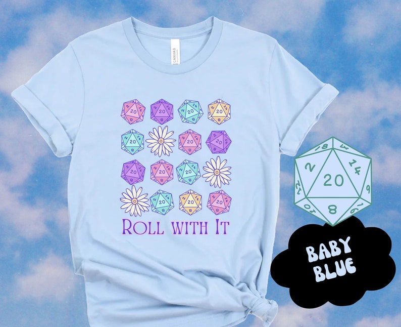 Puede incluir: Una camiseta azul claro con un dise&ntilde;o de varios dados de veinte caras de color pastel y margaritas blancas. El texto "ROLL WITH IT" est&aacute; impreso debajo del dise&ntilde;o. Un solo dado de veinte caras se muestra en la esquina superior derecha de la imagen con el texto "BABY BLUE" en un bocadillo.