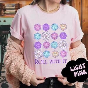 Puede incluir: Una camiseta rosa claro con un dise&ntilde;o gr&aacute;fico de dados coloridos y flores. El texto "ROLL WITH IT" est&aacute; impreso en la camiseta.