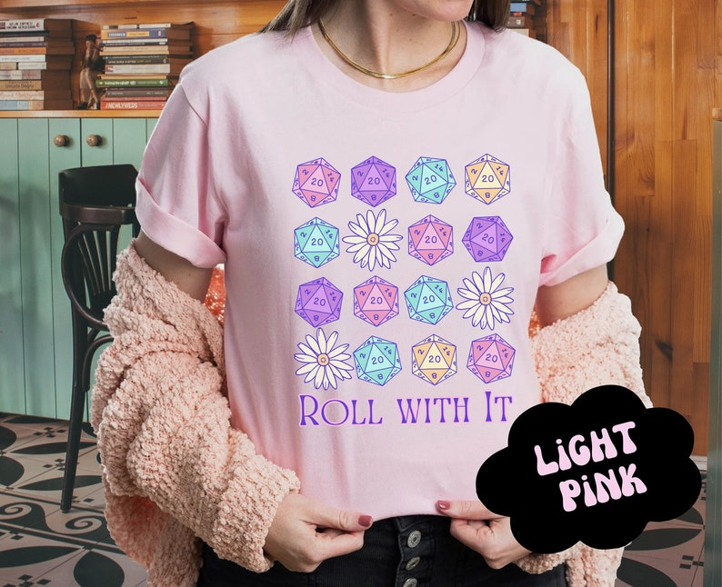 Puede incluir: Una camiseta rosa claro con un dise&ntilde;o gr&aacute;fico de dados coloridos y flores. El texto "ROLL WITH IT" est&aacute; impreso en la camiseta.
