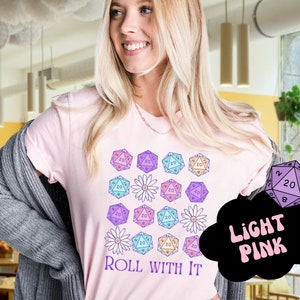 Puede incluir: Una camiseta rosa con un dise&ntilde;o de dados coloridos y flores. El texto "ROLL WITH IT" est&aacute; impreso en la camiseta. La camiseta es rosa claro.