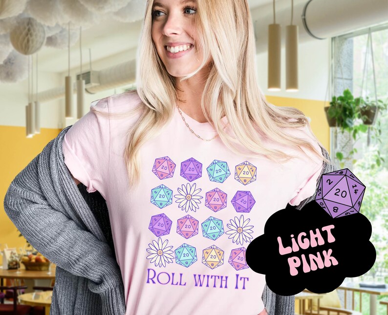 Puede incluir: Una camiseta rosa con un dise&ntilde;o de dados coloridos y flores. El texto "ROLL WITH IT" est&aacute; impreso en la camiseta. La camiseta es rosa claro.