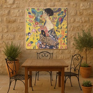 May include: A framed print of a Gustav Klimt painting, featuring a woman in a patterned dress against a yellow floral background. The artwork is displayed above a wooden table and wrought iron chairs on a stone patio.