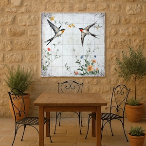 May include: A square wall art piece depicting two swallows in flight, with a floral and botanical design. The artwork is mounted on a stone wall above a wooden table and wrought iron chairs, creating an outdoor dining scene.