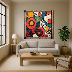 May include: Abstract art piece with a wooden frame. The artwork features bold shapes and colors, including red circles, yellow ovals, and blue and green curved forms. The piece is mounted above a light-colored sofa in a living room setting.