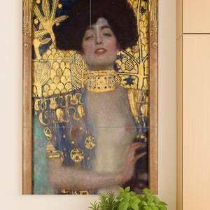 May include: A framed print of a Gustav Klimt painting. The artwork features a woman with dark hair and a gold necklace, set against a backdrop of gold and blue decorative elements. The painting is displayed on a white wall.
