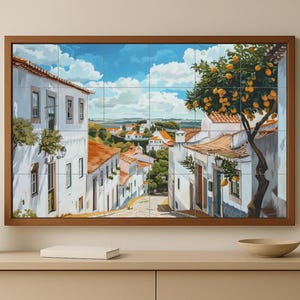 May include: A framed artwork depicting a European village scene. The painting shows white buildings with orange tile roofs, a tree with yellow fruit, and a blue sky with fluffy clouds. The artwork is displayed on a light-colored cabinet.