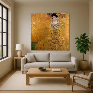 May include: A large framed print of Gustav Klimt's "Portrait of Adele Bloch-Bauer I" hangs above a light-colored sofa. The painting features a woman in a gold dress with intricate patterns. The room has a neutral color palette with a wooden coffee table and a potted plant.