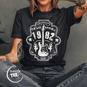 May include: Black t-shirt with a white graphic featuring a guitar, flames, and the text "Vintage Original 1982." The design is framed by a decorative border with the word "Heartbreaker" at the top. The shirt has a crew neck.