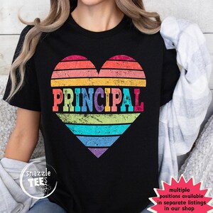 May include: Black t-shirt featuring a rainbow-striped heart design with the word "PRINCIPAL" in bold, colorful letters. The shirt has a crew neck and short sleeves. The design is a retro style.