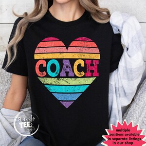 May include: Black t-shirt featuring a heart-shaped design with a rainbow striped pattern. The word "COACH" is printed in colorful letters across the center of the heart. The shirt has a crew neck and short sleeves.