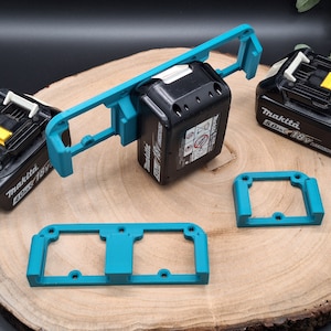Könnte beinhalten: Eine Sammlung schwarzer Makita 18V-Akkupacks und türkisfarbener 3D-gedruckter Batteriehalter. Die Halter sind zur Sicherung der Batterien konzipiert. Das Bild ist auf einer Holzoberfläche platziert.