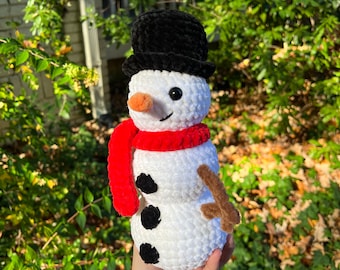 May include: A white crocheted snowman with a black top hat, red scarf, and brown twig arms. The snowman has black eyes, a carrot nose, and three black buttons.