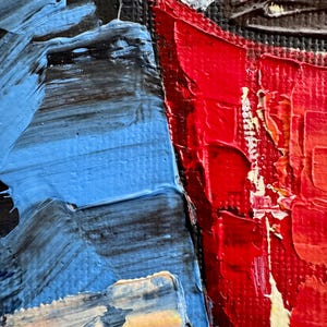 May include: Close-up of a red painted cup with a white rim and a black interior. The cup is painted with thick strokes of red paint, giving it a textured appearance. The background is a blue and black abstract painting.