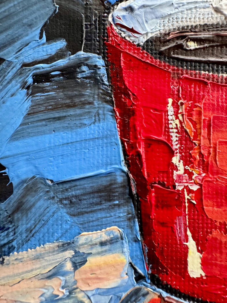 May include: Close-up of a red painted cup with a white rim and a black interior. The cup is painted with thick strokes of red paint, giving it a textured appearance. The background is a blue and black abstract painting.