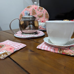 May include: A tea set on a wooden table. The set includes a glass teapot with a silver handle, a white teacup on a saucer, and two coasters. A pink tea cosy with gingerbread house and tree designs is in the background. The coasters and cosy have a pink gingham pattern.