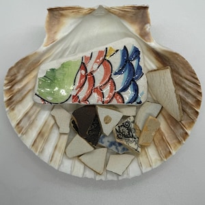 May include: A seashell with a variety of colorful ceramic shards inside. The largest shard is a blue, green, red, and white fish design.