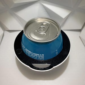 May include: A blue beverage can with a silver top sits inside a white and black holder. The can has text that reads "WARNING: (1) ACCORDING TO THE WOMEN SHOULD NOT DRINK ALCOHOLIC BEVERAGES BECAUSE OF THE RISK". The holder is on a white surface.