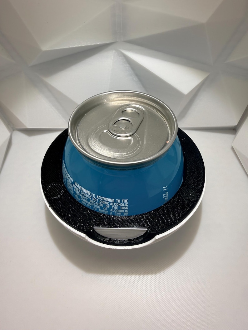 May include: A blue beverage can with a silver top sits inside a white and black holder. The can has text that reads "WARNING: (1) ACCORDING TO THE WOMEN SHOULD NOT DRINK ALCOHOLIC BEVERAGES BECAUSE OF THE RISK". The holder is on a white surface.