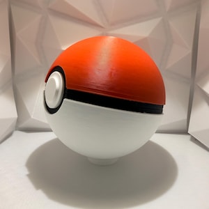 May include: A 3D-printed Pok&eacute; Ball, a spherical device used in the Pok&eacute;mon franchise. The top half is bright orange, the bottom half is white, and a black band separates the two. A white button is on the front.