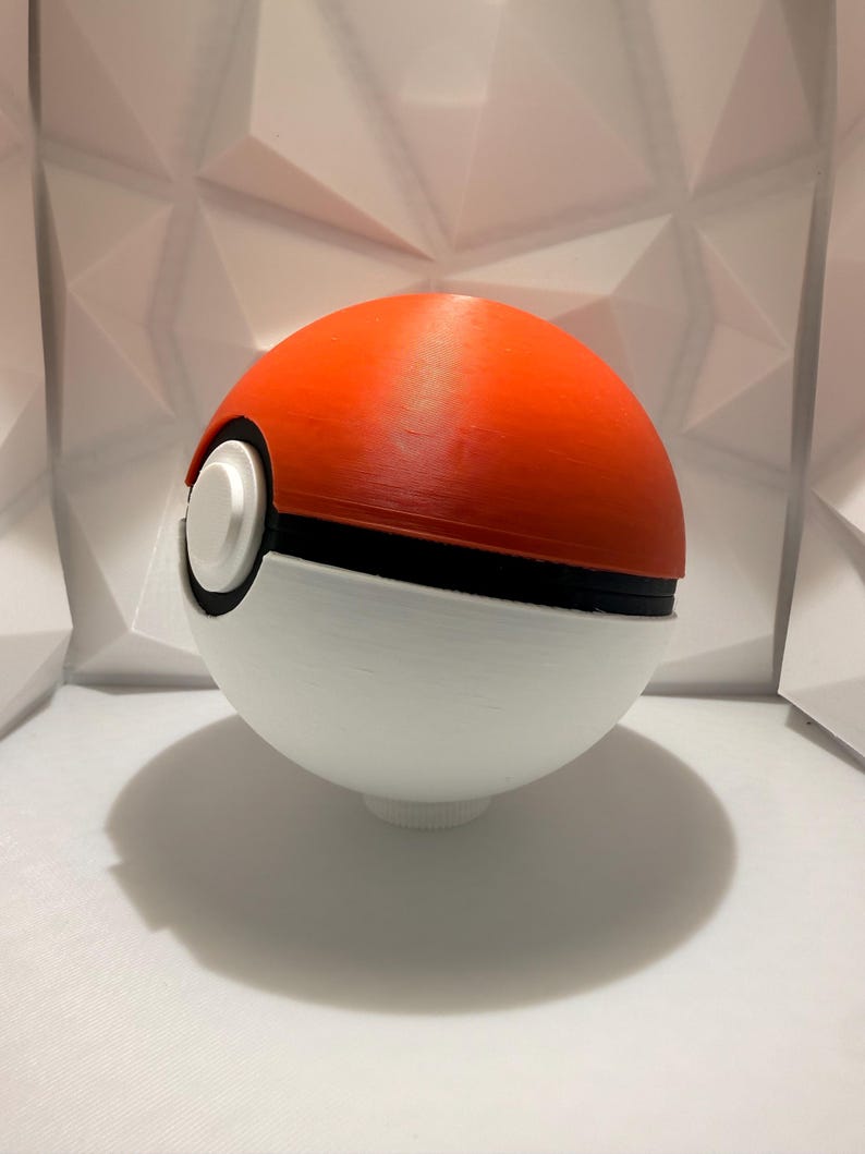 May include: A 3D-printed Pok&eacute; Ball, a spherical device used in the Pok&eacute;mon franchise. The top half is bright orange, the bottom half is white, and a black band separates the two. A white button is on the front.