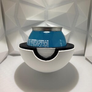 May include: A blue beverage can with silver top, held in a white and black spherical holder. The holder has a circular opening. Text on the can warns against alcohol consumption during pregnancy. The background is a white, geometric pattern.