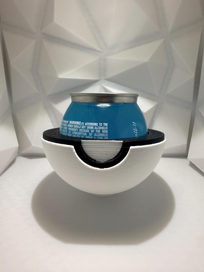 May include: A blue beverage can with silver top, held in a white and black spherical holder. The holder has a circular opening. Text on the can warns against alcohol consumption during pregnancy. The background is a white, geometric pattern.