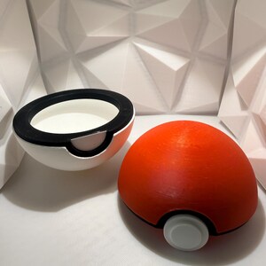 May include: A red and white spherical container, resembling a cartoon Pokeball, is shown open and closed. The container is split in half, with a black rim and a white interior. The other half is a solid red sphere with a white button.